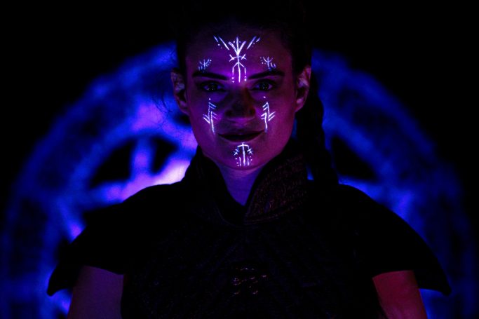 Femme avec des motifs lumineux sur le visage, devant un arrière-plan sombre et coloré.