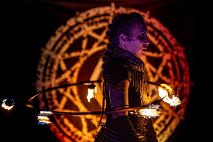 Artiste de feu performe devant un fond avec un motif magique. Lumières vives et expression intense.