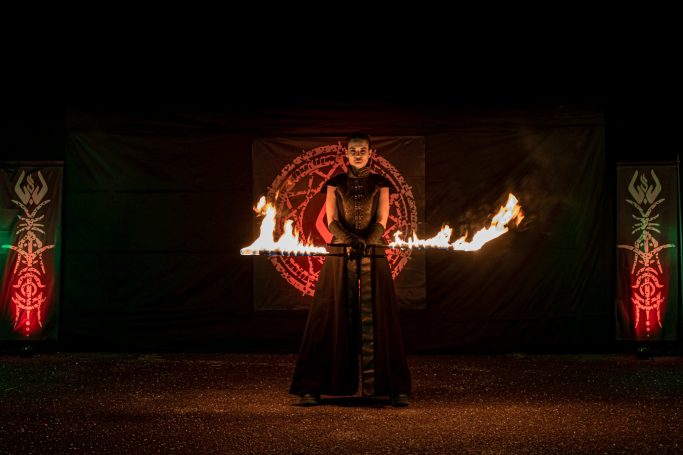 Danseur de feu en costume noir, tenant des épées de feu devant un symbole magique.