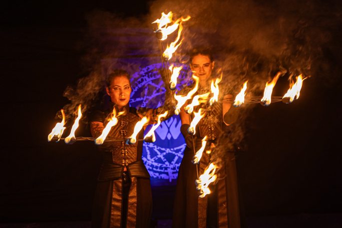Deux performeuses en costumes sombres manipulant des éventails enflammées sur fond bleu.